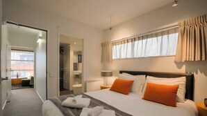Habitación de hotel con una cama grande, dos almohadas, una mesita de noche con lámpara, una ventana con cortinas y un baño visible a través de una puerta abierta.