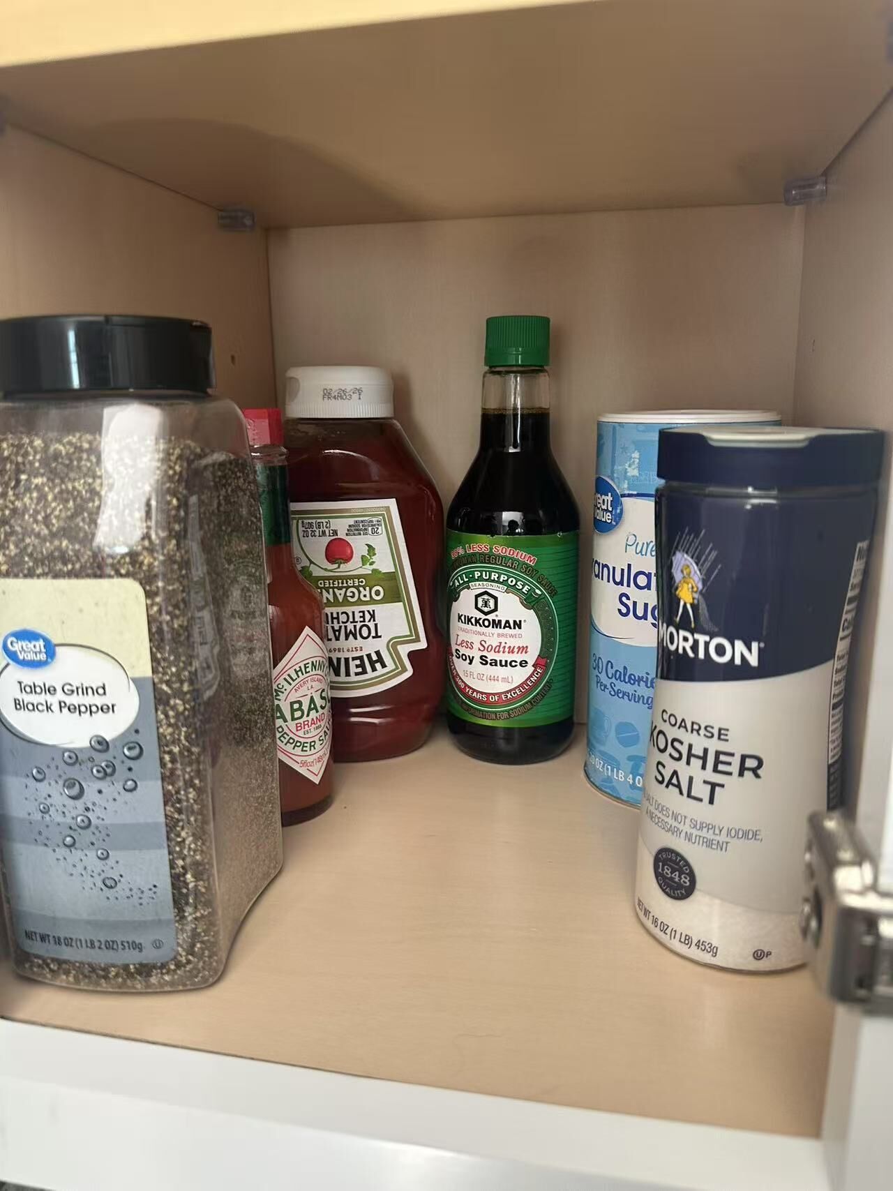A kitchen cabinet with various food items including a jar of black pepper, ketchup, soy sauce, and kosher salt.