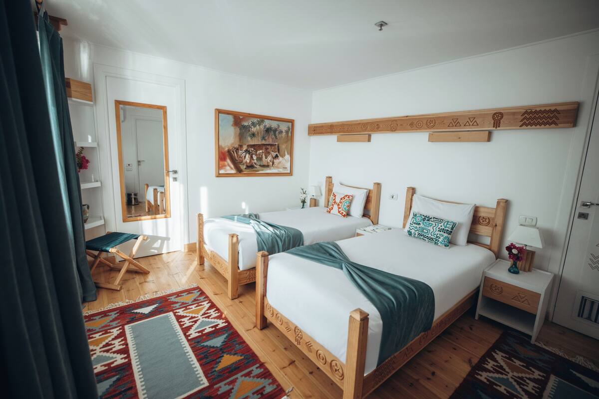 Une chambre d’hôtel avec deux lits en bois, un grand miroir, un tableau et un tapis coloré.