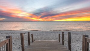 Ein hölzerner Steg führt zu einem Strand, im Hintergrund ist ein Sonnenuntergang zu sehen.