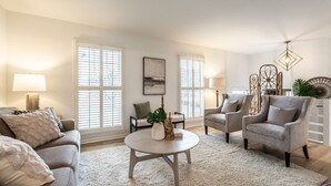 A living room with a sofa, two armchairs, a coffee table, and a lamp. There are windows with blinds, a painting on the wall, and a decorative light fixture. - Groovy Cary Crib - Pickleball, Pets & Plug-In 4 EV (Cary)