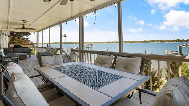 Una terraza techada al aire libre con una mesa, sillas, ventilador de techo y vista a un lago.