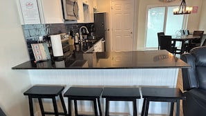 A modern kitchen with a central island, bar stools, and a dining area with a table and chairs. - Sunset By The Sea 3054 - FMB (Fort Myers Beach)