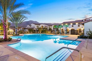 A swimming pool with lounge chairs and palm trees, surrounded by buildings with a mountain backdrop.