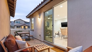 Eine Terrasse mit einer Couch, einem Holztisch und Stühlen, sowie eine Glastür, die zu einem Innenbereich mit Fernseher und Sitzgelegenheiten führt.