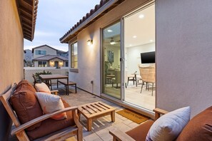Een terras met een bank, houten tafel en stoelen, een schuifpui naar een binnenruimte met een televisie en zitplaatsen.