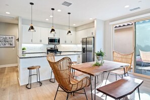 A modern kitchen with white cabinets, a central island, pendant lighting, and a dining area with wicker chairs. - Villa Solara: EV Charger, Pool, Spa & PGA West Gym (La Quinta)