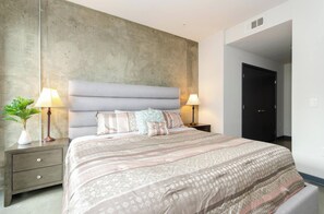 A bedroom with a large bed, two bedside tables with lamps, a potted plant, and a dark door. - Lovely 3BR Apt w/ Balcony & Rooftop Pool + Views (Los Angeles)
