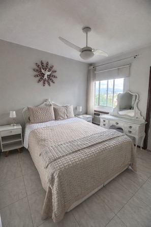Un dormitorio con cama, mesita de noche, cómoda, ventilador de techo, ventana con cortinas y un adorno en la pared.