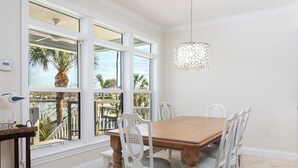 A dining area with a wooden table and white chairs, large windows with a view of palm trees and a balcony, and a hanging light fixture. - Welcome to Bluefin – Your Waterfront Getaway! (Treasure Island)