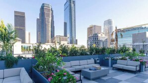 A rooftop patio with outdoor seating, surrounded by greenery and overlooking a cityscape with modern buildings. - City Skyline Views 3-Bed High-rise Apartment w/Pool, Spa & Rooftop Views (Los Angeles)
