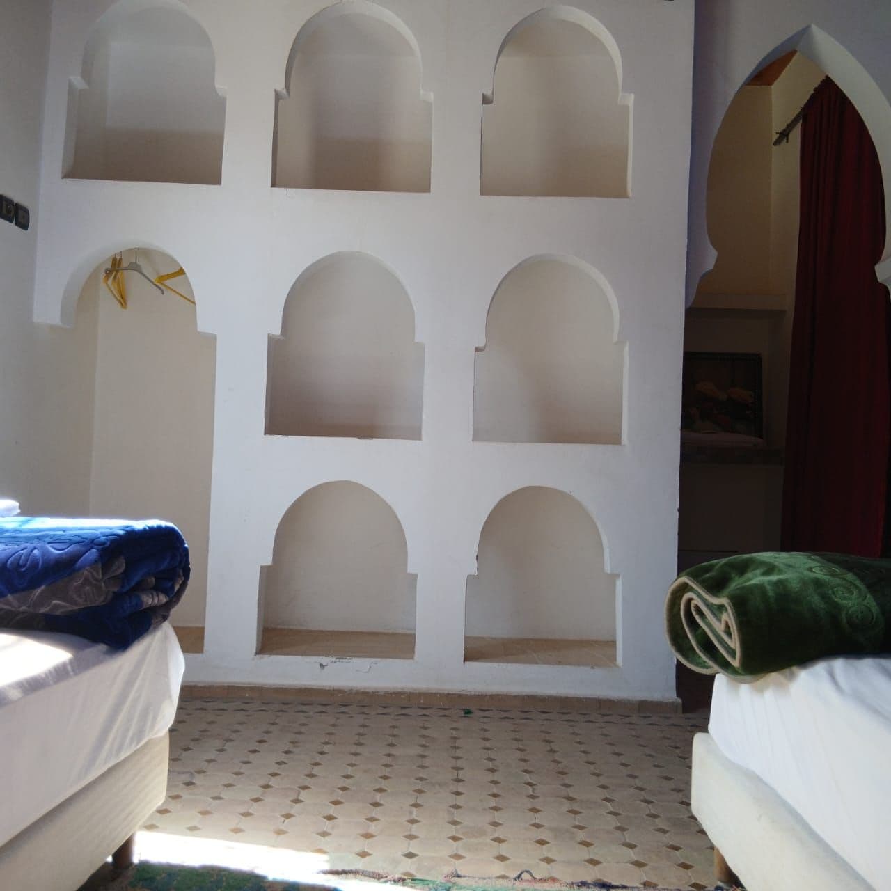 A room with a patterned floor, a bed with a blue blanket, and a wall with multiple arched niches.