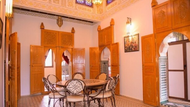 Un interior tradicional marroquí con una mesa redonda para comer rodeada de sillas, puertas de madera ornamentadas y baldosas de suelo con diseños.