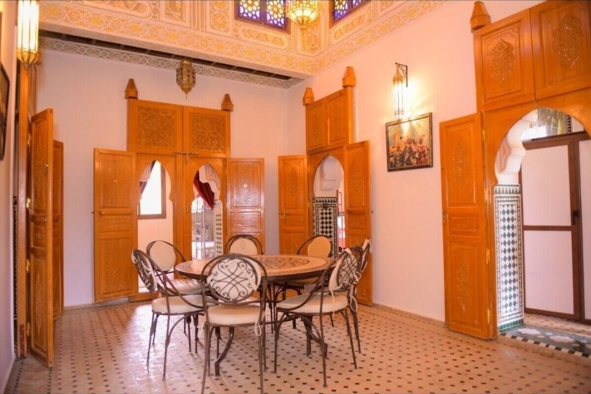 Un intérieur marocain traditionnel avec une table à manger ronde entourée de chaises, des portes en bois richement décorées et des carreaux de sol aux motifs variés.