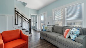 Una sala de estar con un sofá gris, un sillón naranja y cojines estampados. Una escalera con barandilla negra conduce a otra habitación.