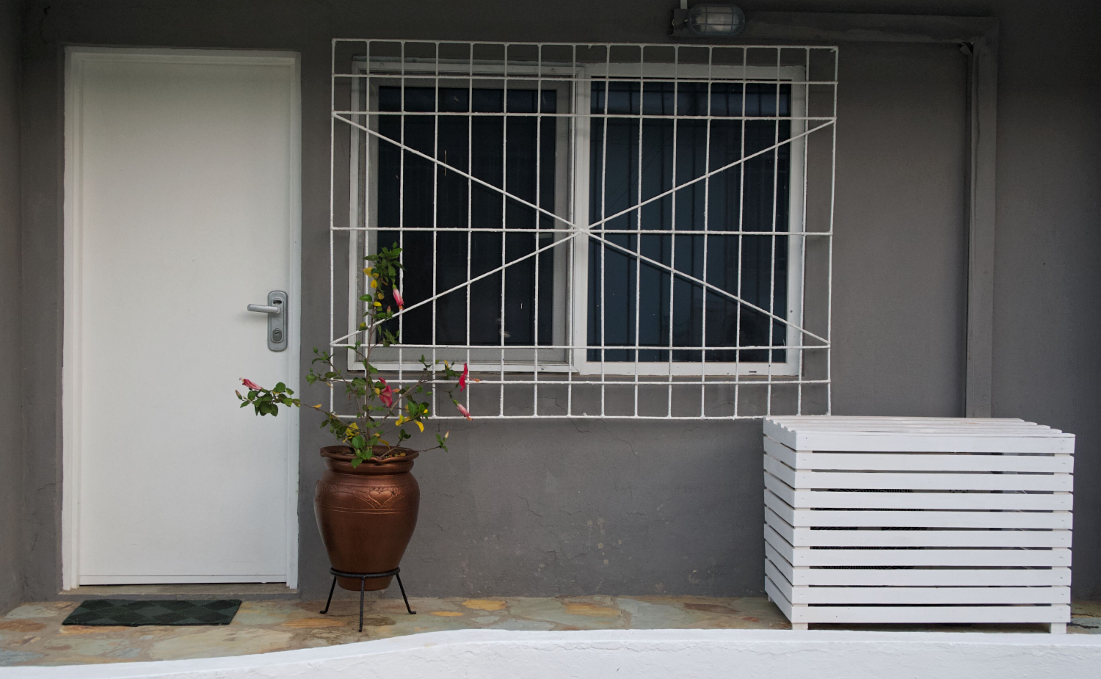 Una puerta, una ventana con una celosía metálica, una planta en maceta y una pila de objetos blancos.