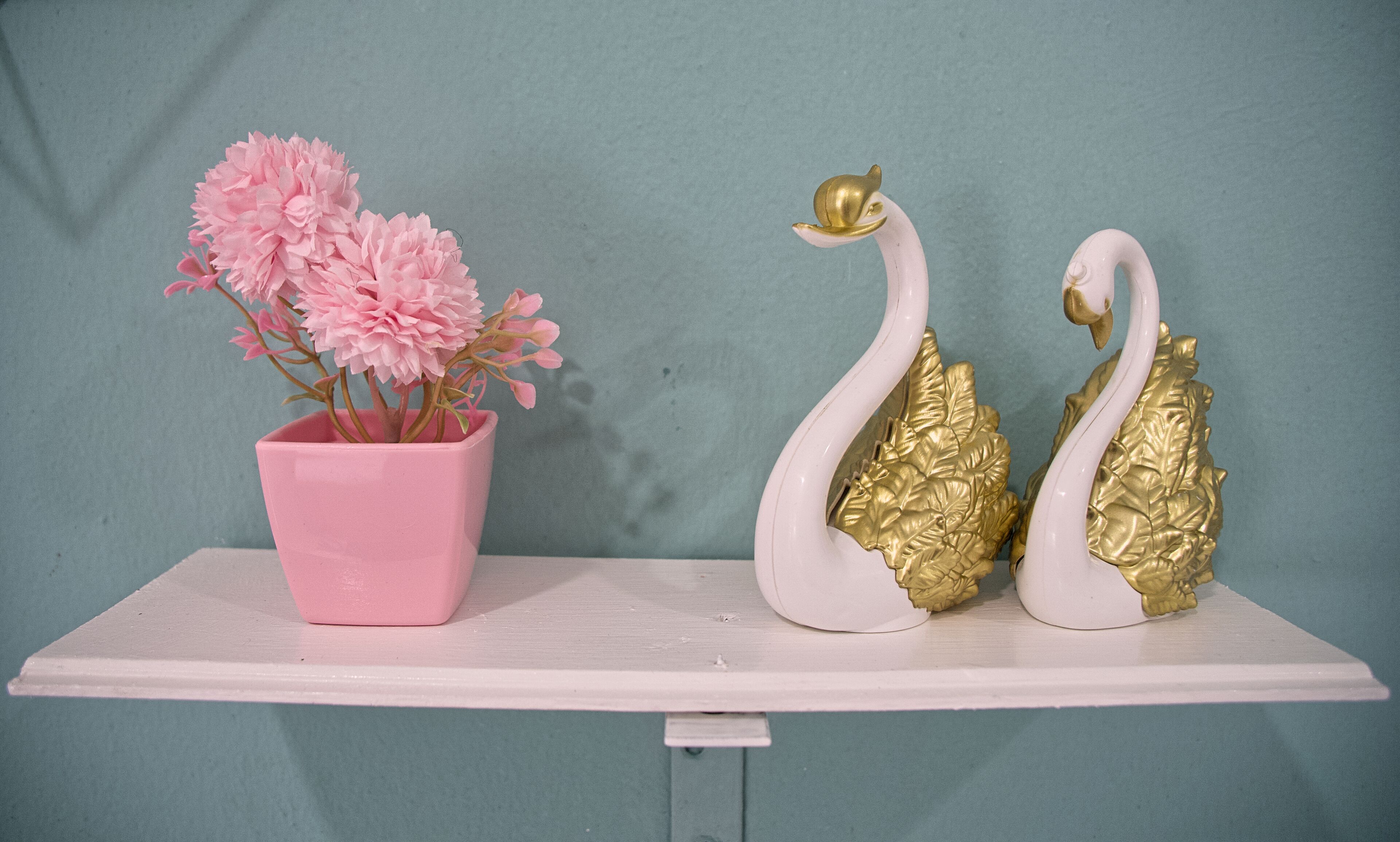 En vit hylla med en rosa vas som innehåller rosa blommor, och två vita svanstatyetter med guldfärgade detaljer.