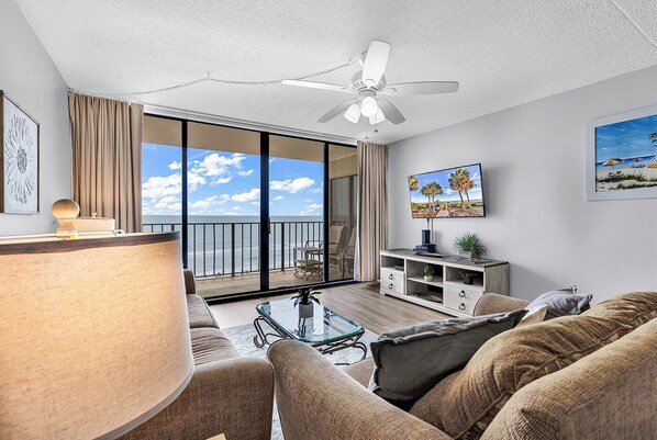 A modern living room with a balcony, a ceiling fan, a TV, and a large window with a view of the ocean. - Horizon East 303 - Oceanfront in Garden City Beach (Garden City)