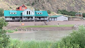 A large house with a green roof and a red roof section, situated near a river and a rocky hillside.