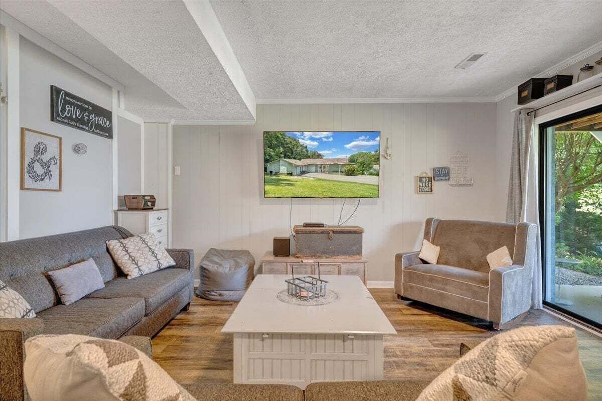 Ein Wohnzimmer mit einem großen Fernseher, zwei Sofas, einem Couchtisch und Blick auf ein Haus durch ein Fenster.