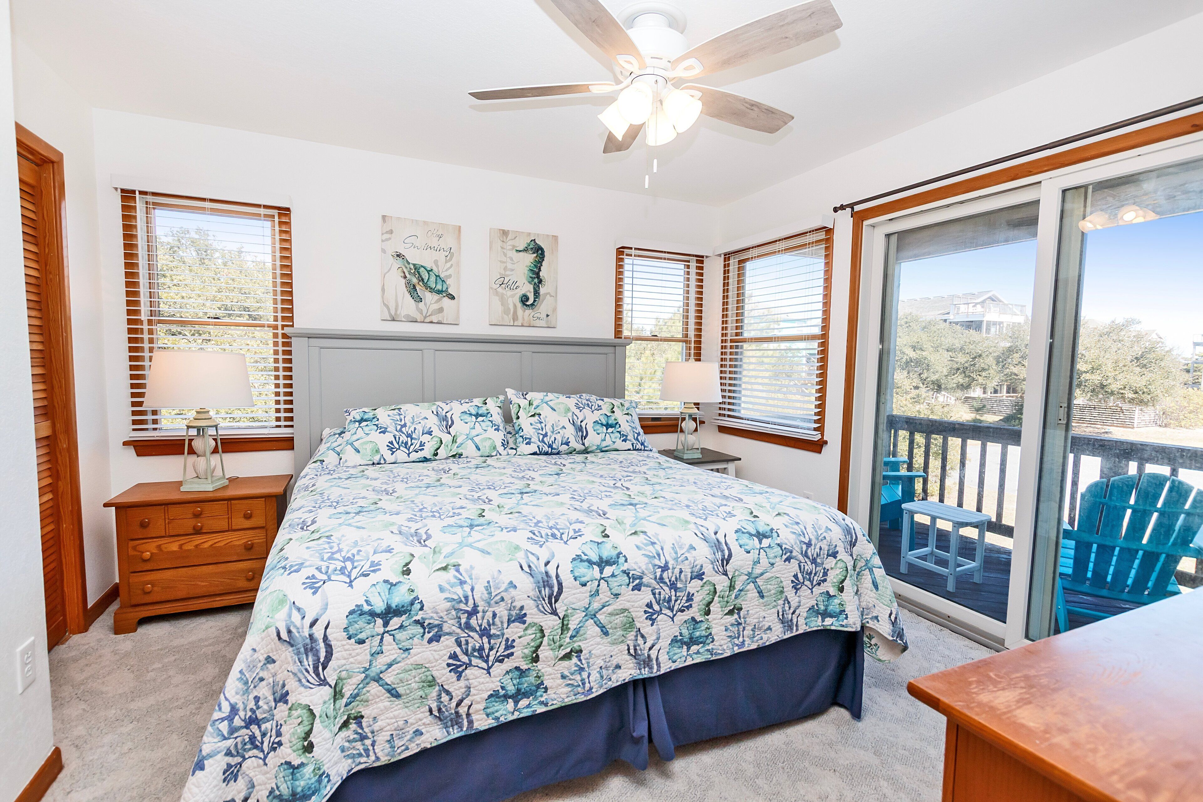 A bedroom with a bed, wooden nightstands, a ceiling fan, and large windows with blinds.