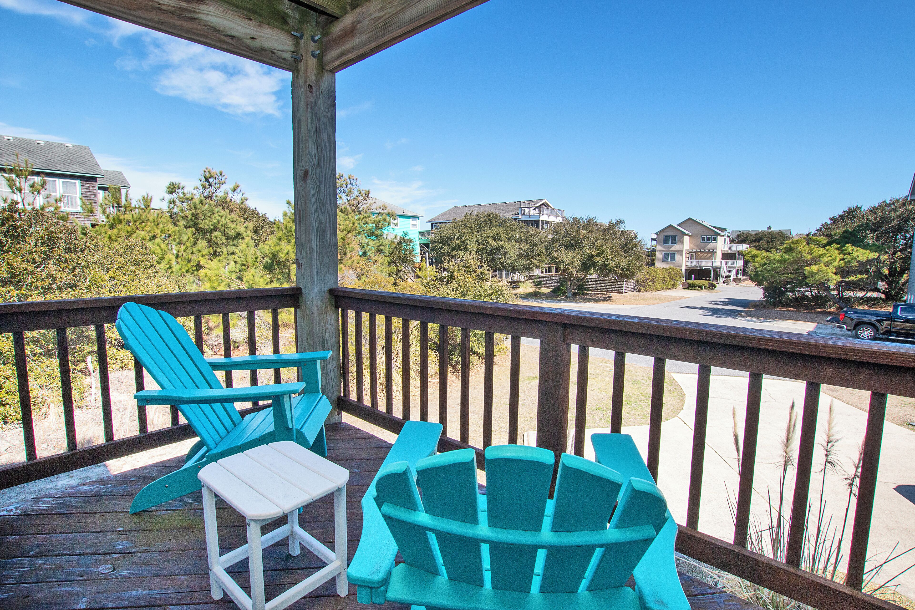 A wooden deck with turquoise Adirondack chairs and a white table, overlooking a residential area with houses and a street.
