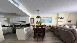 A modern kitchen with white cabinets, a dining area with a wooden table and chairs, and a living area with a brown sofa and a large painting on the wall. - WELCOME TO THE IVORY TOWER AT ARCADIAIN SHORES - 2 BEDROOM - CONDO ON THE OCEAN (Myrtle Beach)