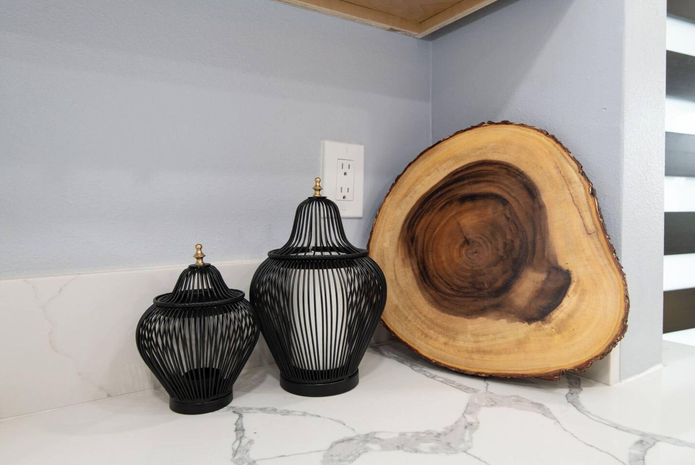 Three black vases with gold accents on a marble countertop, a large wooden slice, and a white electrical outlet on a light blue wall.