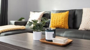 A modern living room with a dark leather sofa, a white potted plant, a wooden tray with a candle and books, and a yellow patterned pillow.
