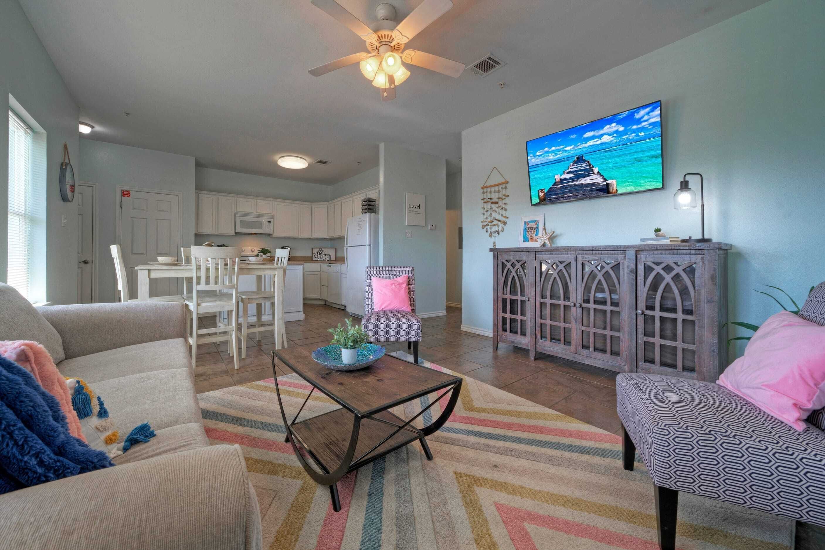A living room with a sofa, a coffee table, and a TV mounted on the wall. There is a kitchen area visible in the background.