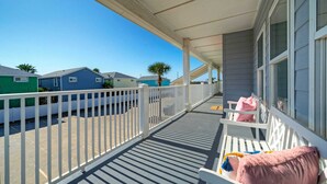 A covered balcony with white railings, a bench with a pink pillow, and a view of neighboring houses.