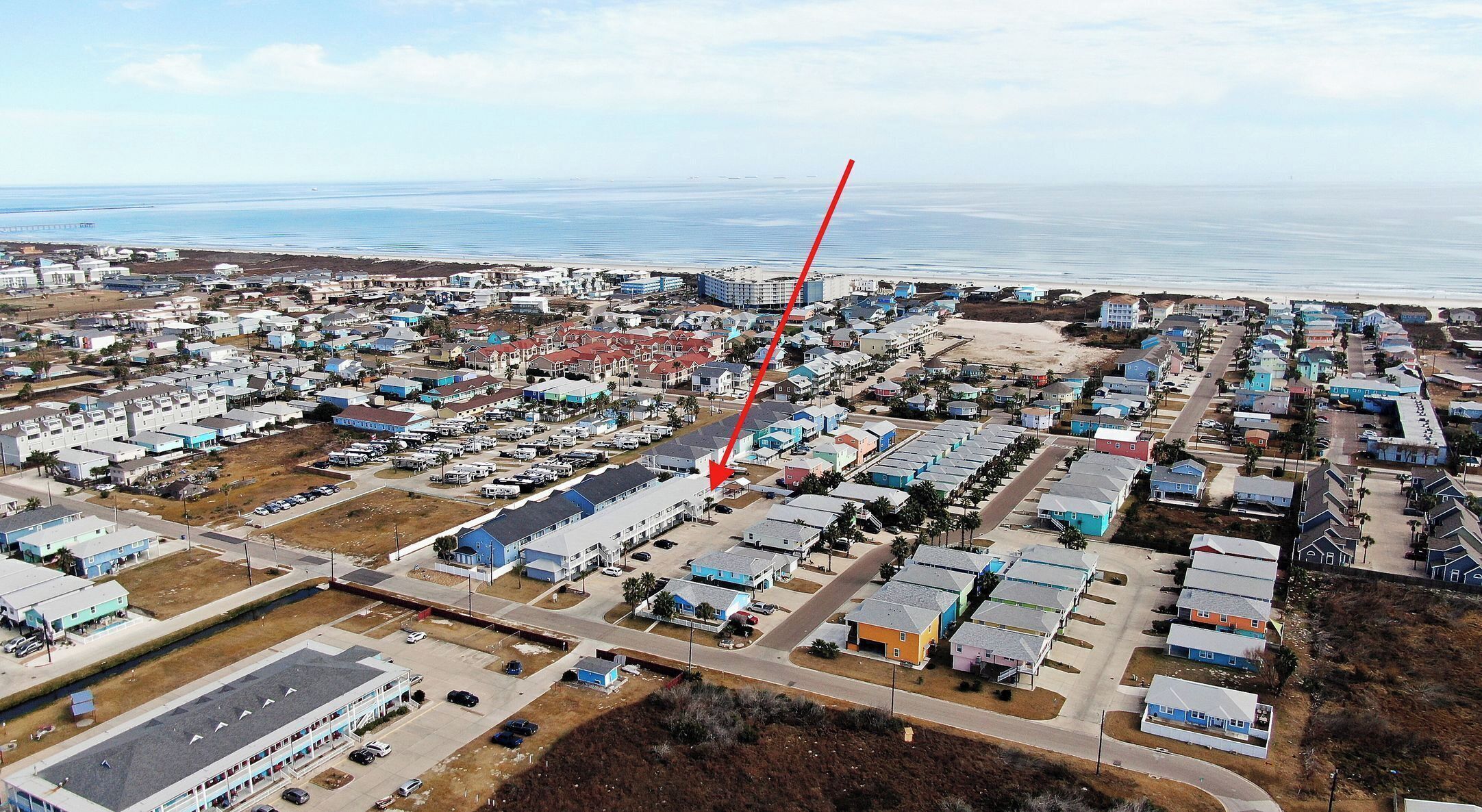 An aerial view of a coastal residential area with a mix of single-family homes and apartment buildings, a beach in the background, and a prominent red arrow indicating a specific location.