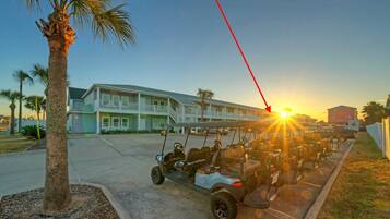 A row of golf carts parked in front of a building with a balcony, palm trees, and a sunset in the background.