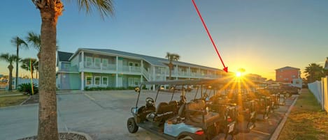 A row of golf carts parked in front of a building with a balcony, palm trees, and a sunset in the background.