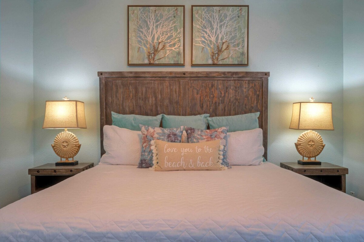 A neatly made bed with a quilted cover, two wall-mounted lamps, and two framed artworks above the bed.