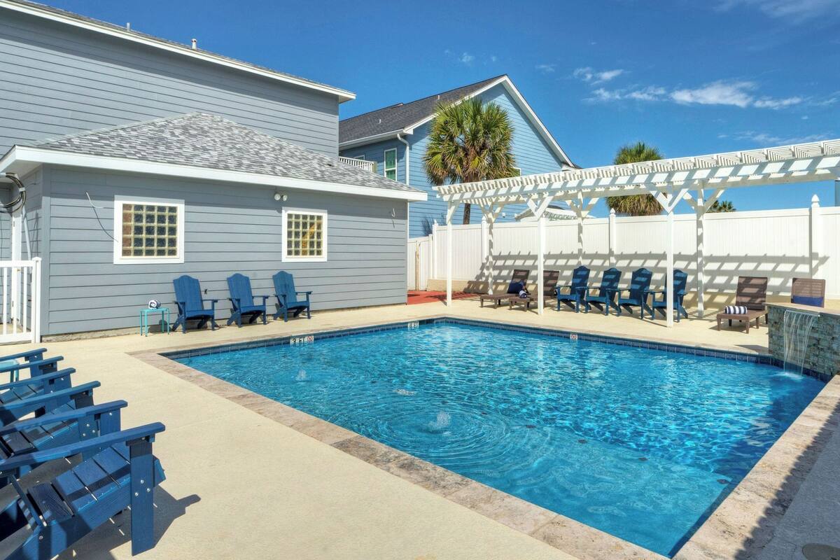 Una piscina con agua azul, rodeada de sillones de descanso y una pérgola.