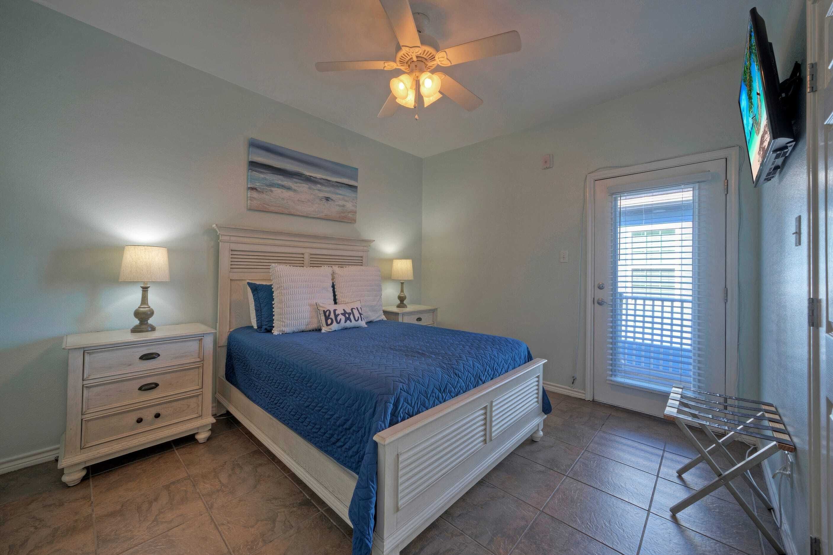 A bedroom with a bed, nightstands, a ceiling fan, a TV, and a door with blinds.