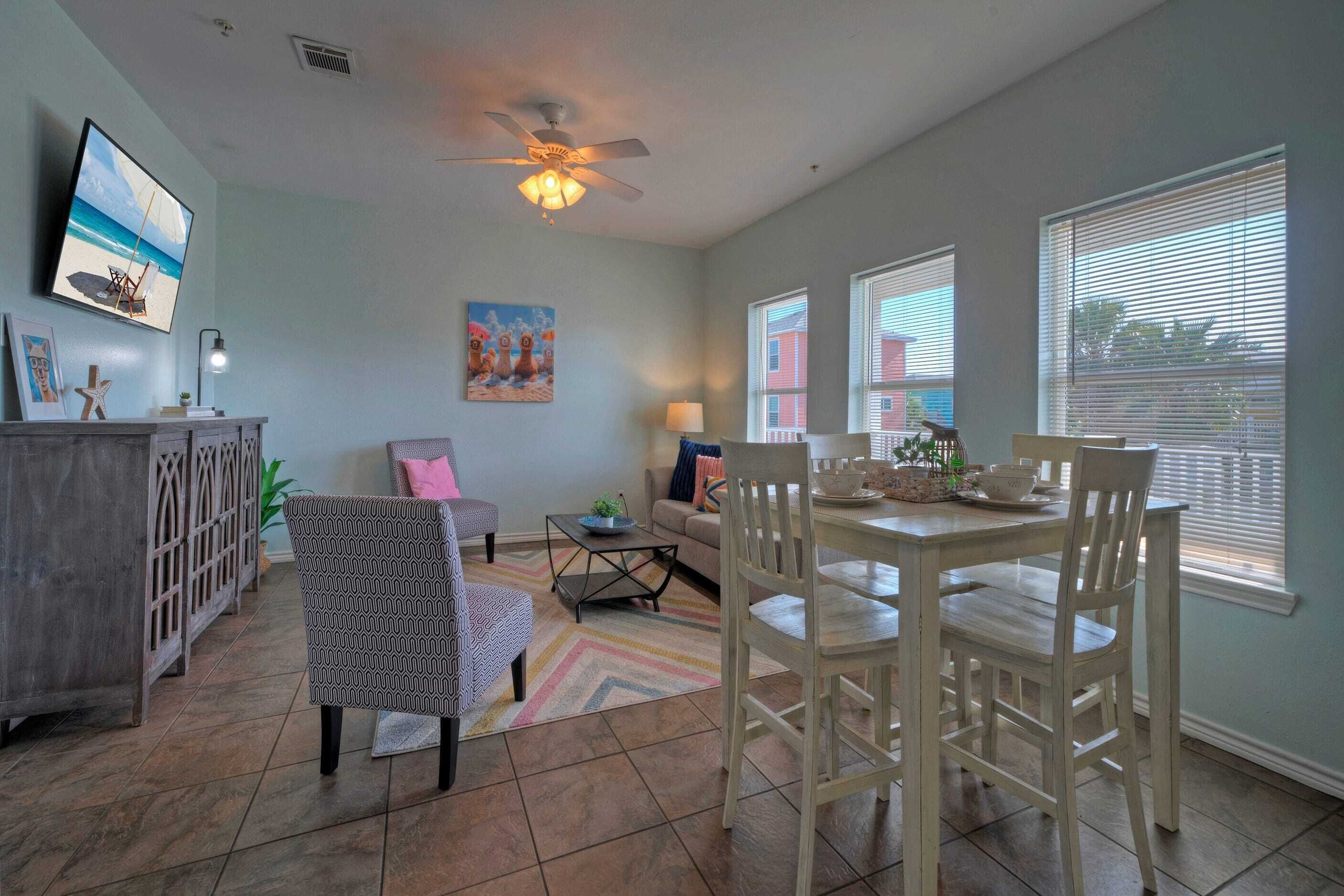 A living room with a dining area, a ceiling fan, a TV mounted on the wall, and a window with blinds.