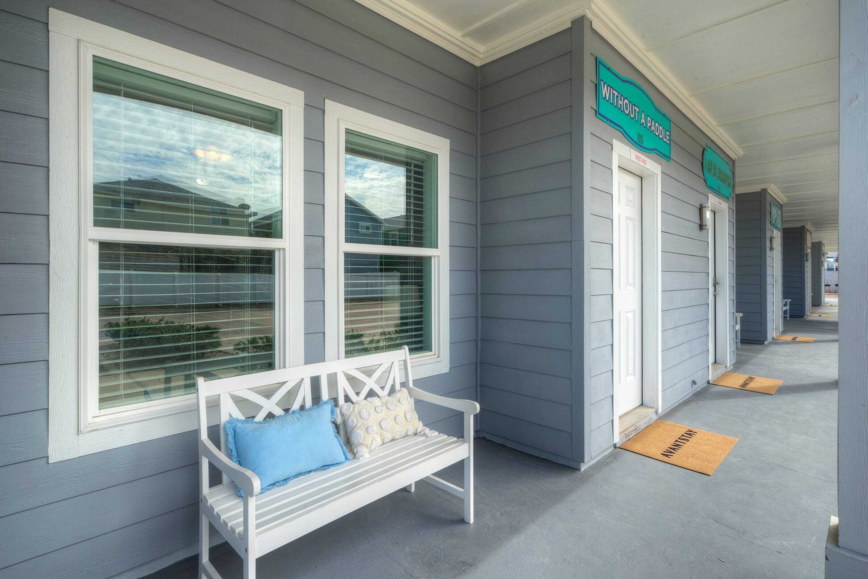 A covered porch with a white bench and a bench with blue cushions. There are two doors with signs above them.