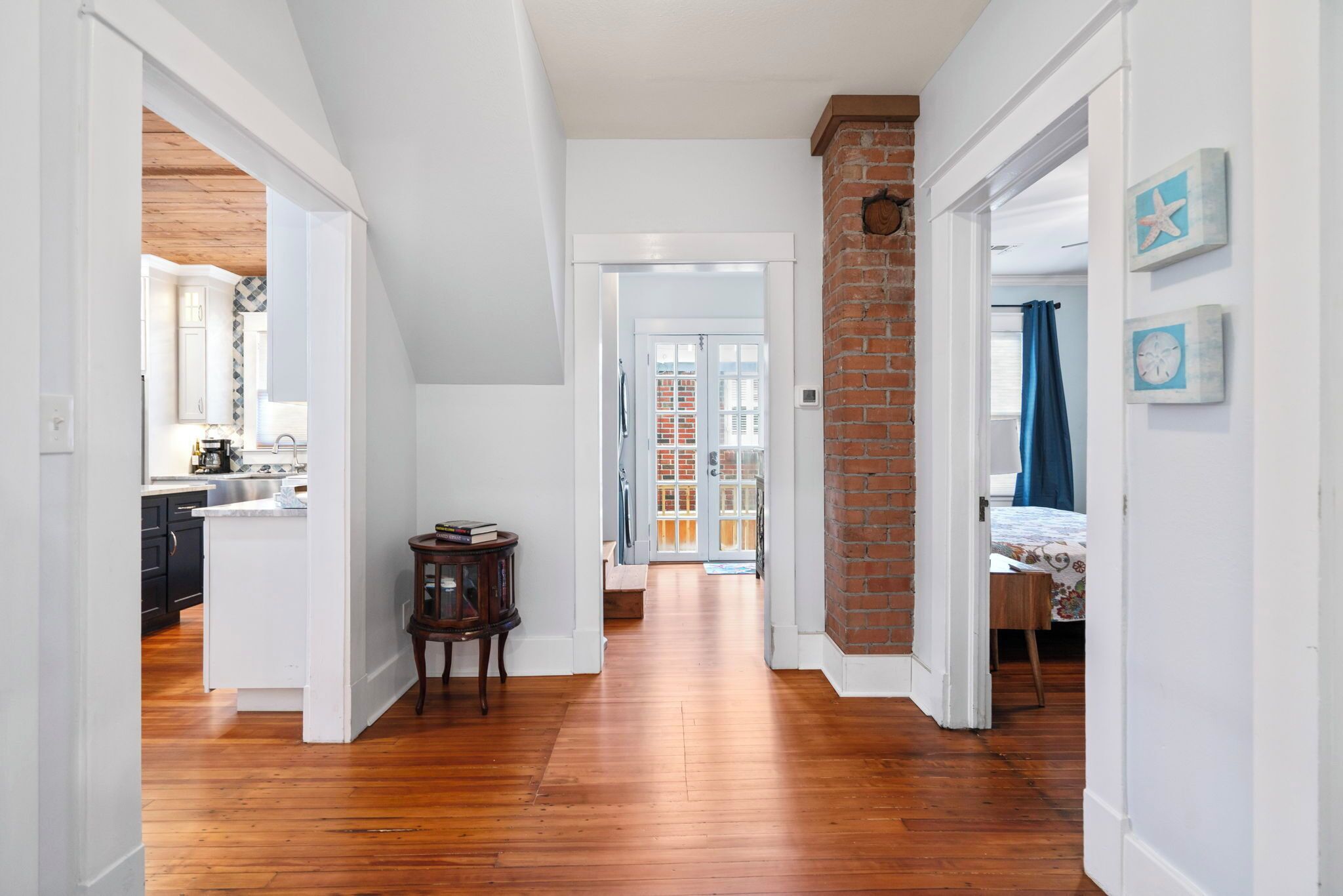 A hallway with wooden flooring, white walls, and a brick accent wall. There is a door leading to a room with a bed and a window with blue curtains.