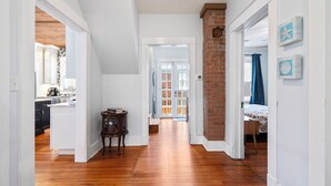 A hallway with wooden flooring, white walls, and a brick accent wall. There is a door leading to a room with a bed and a window with blue curtains.