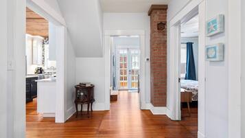 A hallway with wooden flooring, white walls, and a brick accent wall. There is a door leading to a room with a bed and a window with blue curtains.