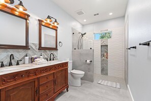 A modern bathroom with a vanity, sink, and toilet. It includes a shower area with a glass door and a view of greenery outside. - Cottage at Kempner Park | Pet-Friendly Modern Home w/ Backyard (Galveston)