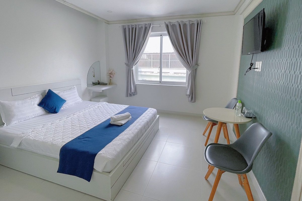 A hotel room with a bed, a small table with chairs, a TV mounted on the wall, and a window with curtains.