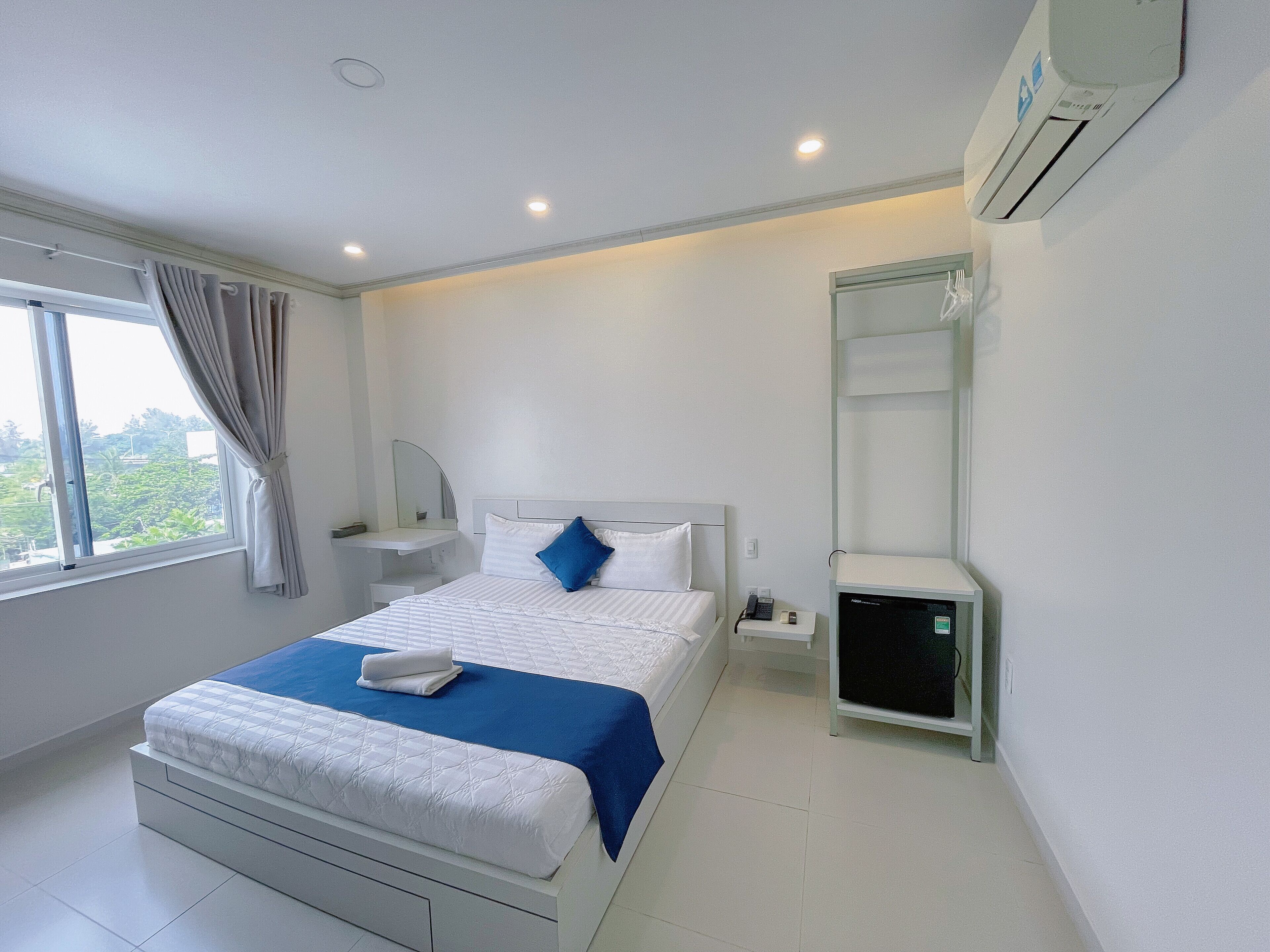 A modern hotel room with a large bed, a bedside table, a small bedside cabinet, a window with curtains, and an air conditioning unit on the wall.