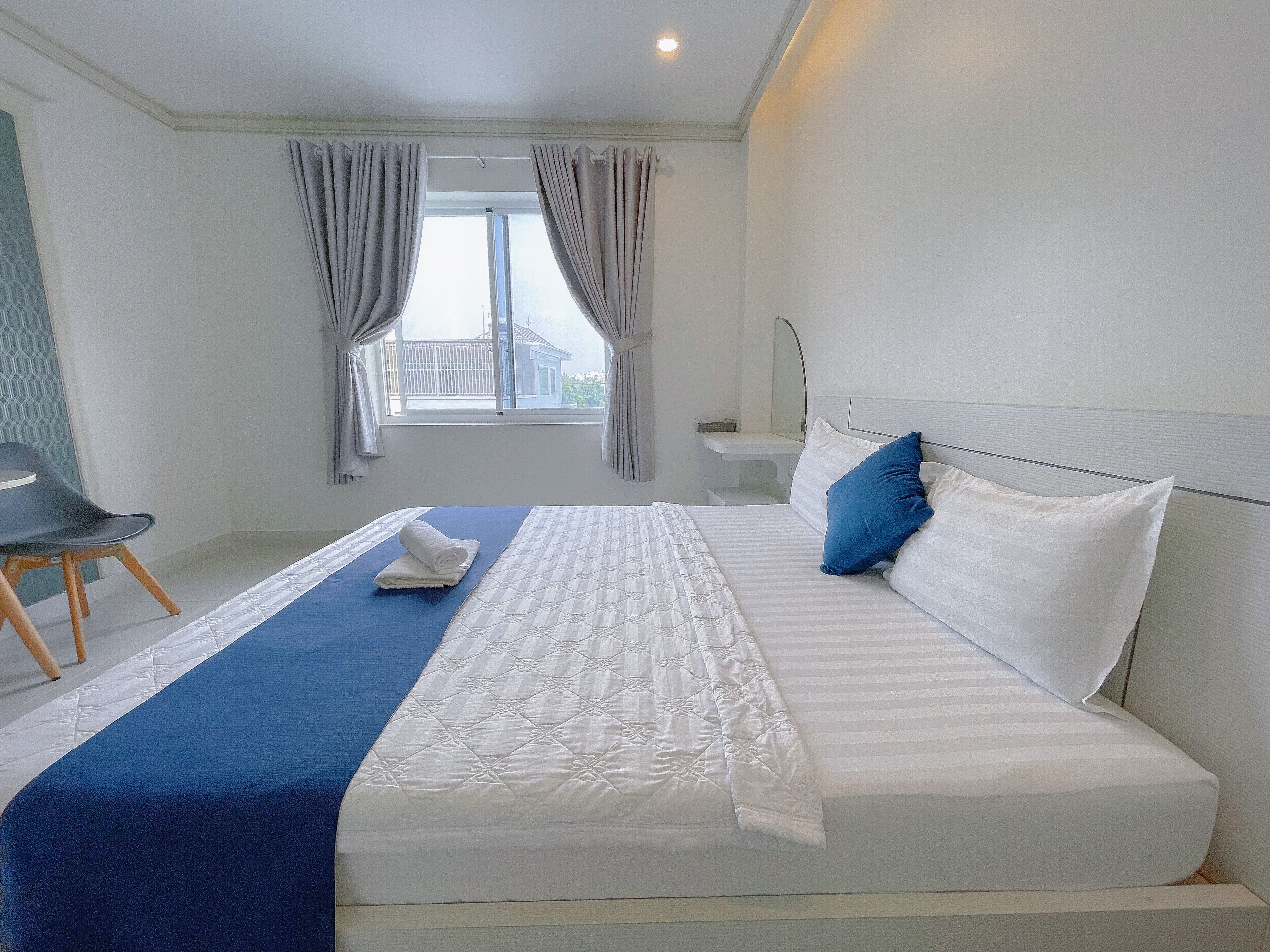 A neatly made bed with a blue bedspread and white pillows, a small bedside table, and a window with curtains.