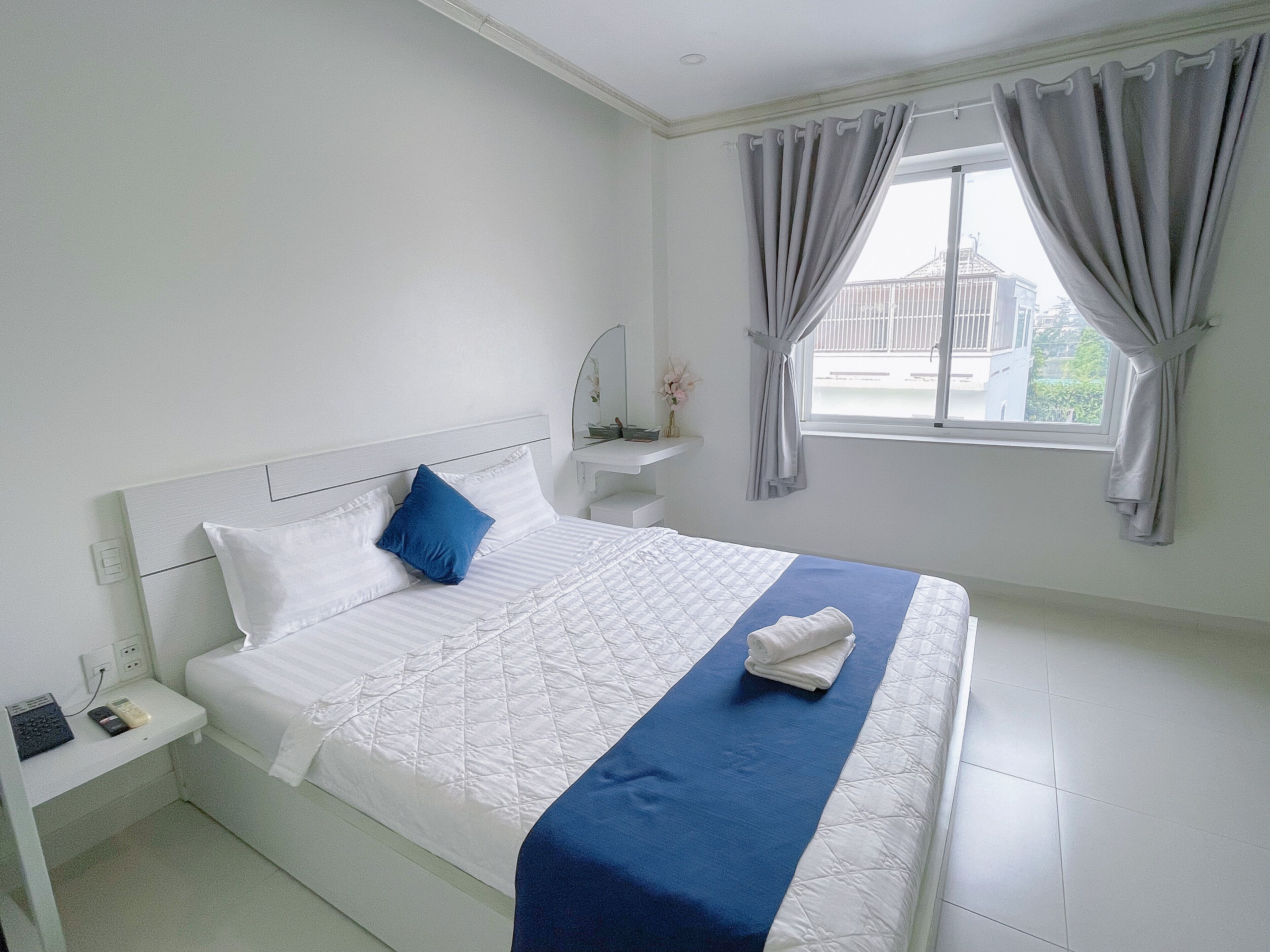A neatly made bed with white and blue bedding, a bedside table with a phone and charger, a small mirror, and a window with curtains.