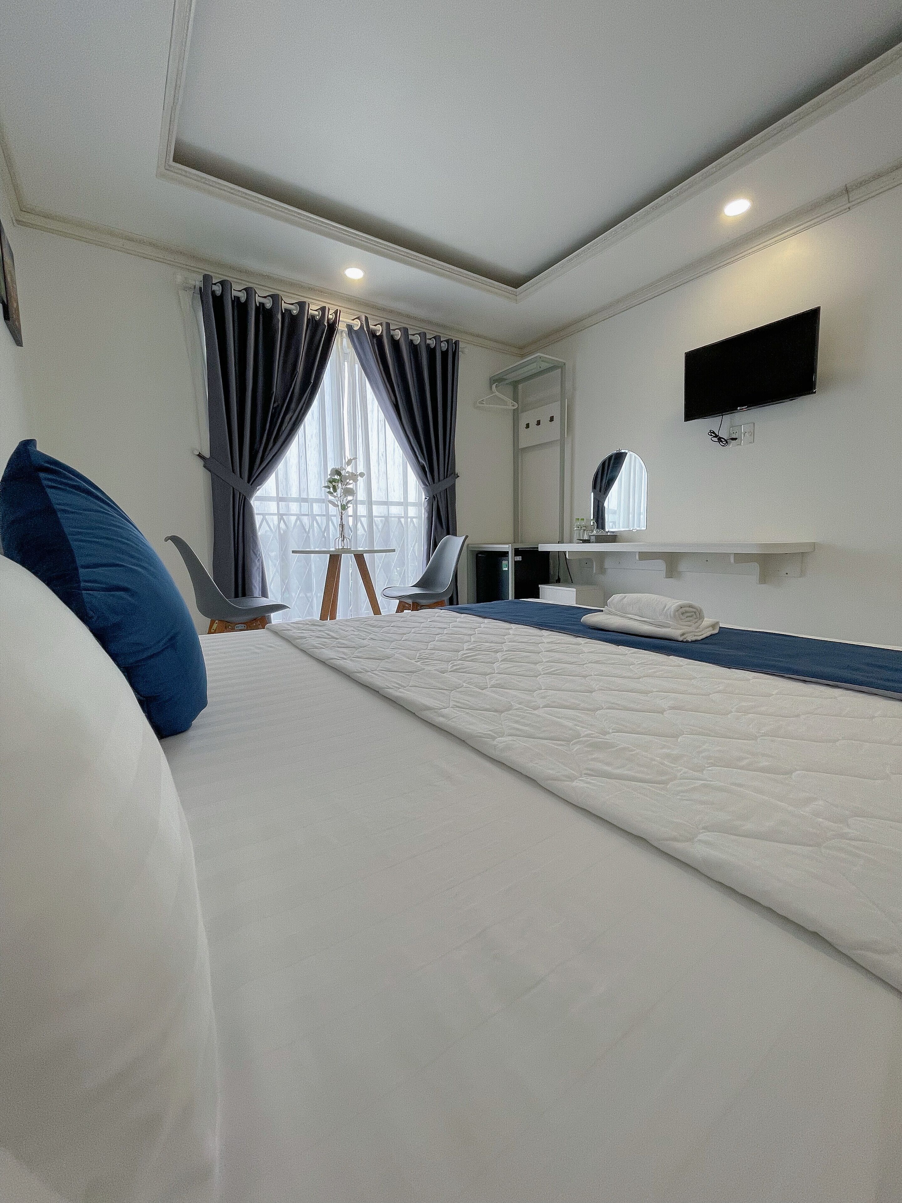 A hotel room with a large bed, a desk with a chair, a flat-screen TV mounted on the wall, and a window with curtains.