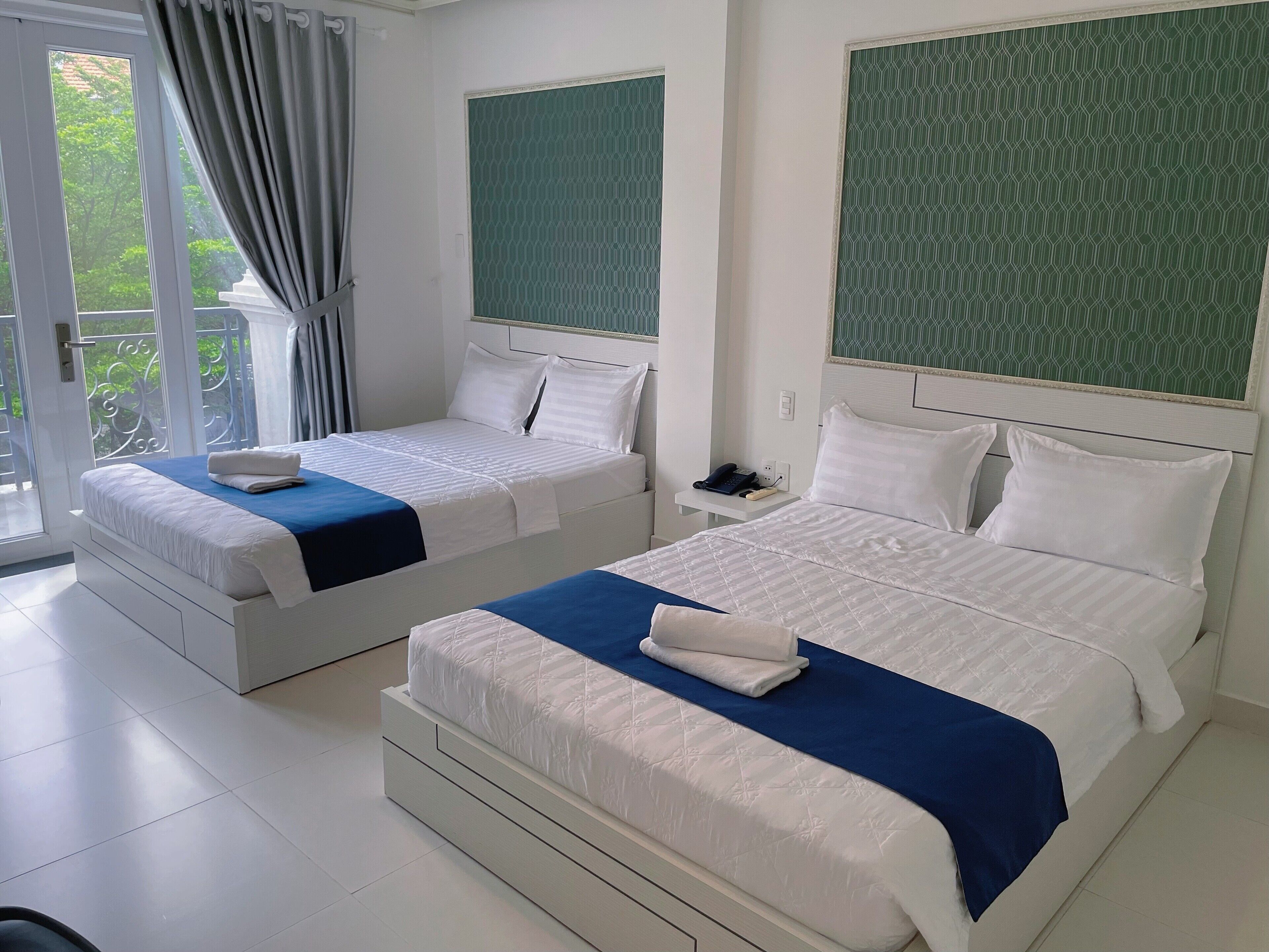 A hotel room with two beds, a large window with curtains, a green patterned wall, and a white tiled floor.