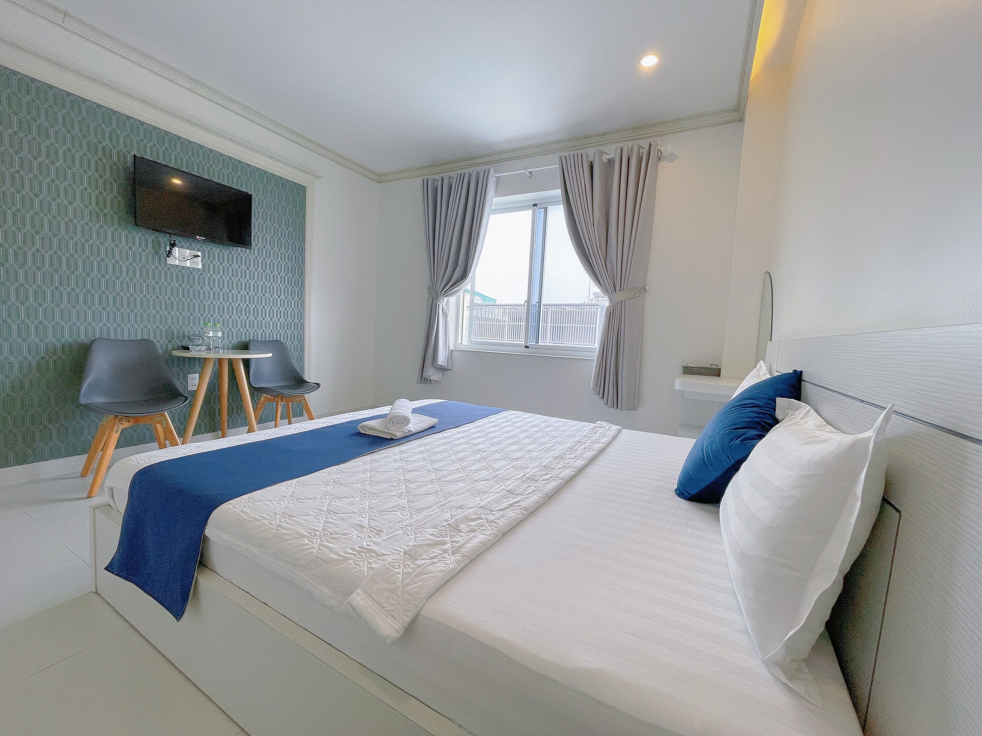 A hotel room with a bed, a TV mounted on the wall, a small table with chairs, and a window with curtains.